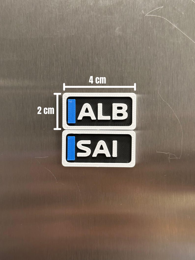 K&ouml;nnte beinhalten: Zwei rechteckige Magnete mit wei&szlig;em Rand, schwarzer Mitte und blauen vertikalen Streifen auf der linken Seite. Der obere Magnet zeigt den wei&szlig;en Text ALB, der untere den wei&szlig;en Text SAI. Jeder Magnet misst 4 cm mal 2 cm.