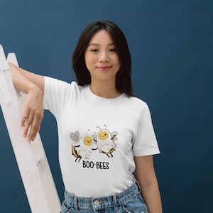May include: White t-shirt with a cartoon illustration of two ghost-bees with smiling faces and wings. The text "BOO-BEES" is printed below the characters in black. The shirt is worn by a person.