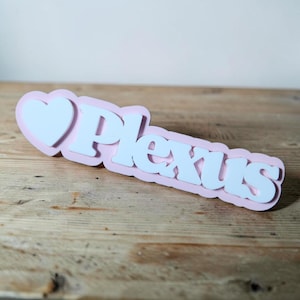 May include: A pink and white Plexus sign with a heart shape. The word "Plexus" is in white, bold letters, and the sign has a layered design. The sign is on a wooden surface.