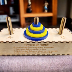 May include: A wooden puzzle game called Towers of Hanoi. The game has three wooden posts labeled A, B, and C. A stack of five yellow and blue rings is on post A. The game instructions are printed on the base of the puzzle: "Move all disks from one tower to another. 1. Only one disk moved at a time. 2. Only the top disk on a stack can be moved. 3. No larger disk on top of a smaller disk."