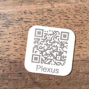 May include: A white, square QR code tag with the word "Plexus" printed in a sans-serif font. The QR code is a complex pattern of black and white squares. The tag is on a wooden surface.