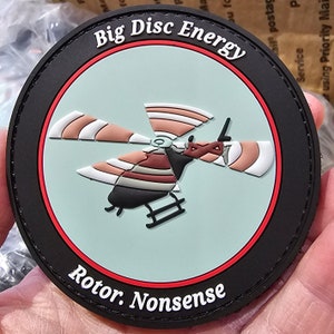 Könnte beinhalten: Ein schwarzer und roter runder Patch mit weißem Text, der "Big Disc Energy" und "Rotor. Nonsense" lautet. Ein Cartoon-Hubschrauber mit einem rot-weiß-braunen gestreiften Rotor befindet sich in der Mitte des Patches.