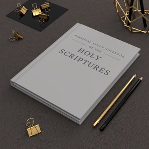 May include: A gray notebook with the title "Personal Study Notebook of the Holy Scriptures". The notebook is on a brown leather surface with two pencils and a gold paperclip.