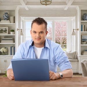 May include: A light blue button-down shirt with a white undershirt, worn by a person sitting at a wooden desk with a blue laptop. The person wears a silver-toned wristwatch on their left wrist while facing forward in a bright, well-lit room.