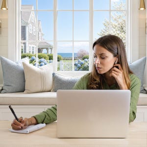 May include: A silver laptop computer sits on a light-colored wooden desk. A person is using a black stylus to write on a small white digital tablet while wearing a green long-sleeved shirt and a white wireless earbud in their right ear.
