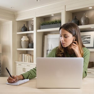 May include: A woman wearing a green shirt is working on a laptop at a desk. She is holding a pen and writing on a notepad. The background includes a built-in cabinet with decorative vases and framed artwork.