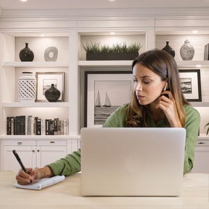 May include: A woman wearing a green shirt is working on a laptop at a desk. She is holding a pen and writing on a notepad. Behind her is a white bookshelf with decorative items, including vases, books, and framed artwork. The background includes a black and white framed picture of sailboats.