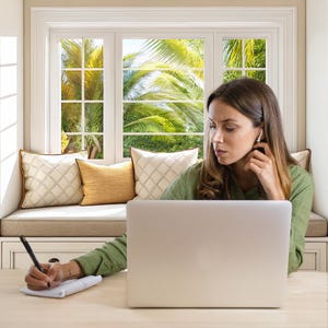 May include: A woman working on a laptop at a desk near a window. She is wearing a green shirt and using a pen to write on a notepad. The window has a view of palm trees and is surrounded by decorative pillows.