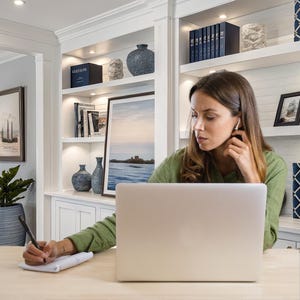 Könnte beinhalten: Eine Frau in einem grünen Hemd arbeitet an einem hellen Holzschreibtisch mit Laptop und Notizblock. Sie hält einen Stift und hat Ohrhörer im Ohr. Hinter ihr befindet sich ein weißes Bücherregal mit Vasen, Büchern und gerahmten Kunstwerken. Der Raum wirkt hell und luftig.