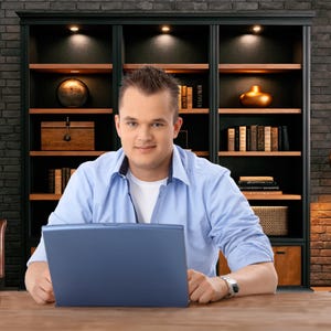 May include: A man in a light blue button-down shirt and a white undershirt sits at a wooden desk, using a blue laptop. Behind him is a dark wooden bookcase with books and decorative items. The background features a brick wall.