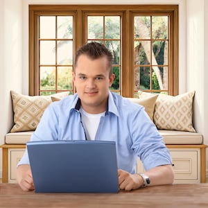 May include: A person seated at a wooden table, working on a blue laptop. The person is wearing a light blue button-down shirt over a white t-shirt. The background includes a window with a view of trees and decorative pillows.
