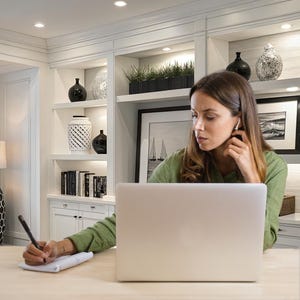 May include: A woman wearing a green shirt is working on a laptop and taking notes. The workspace includes a notebook, pen, and a built-in bookshelf with decorative vases and framed artwork. The room has a neutral color palette.