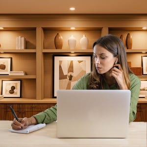 May include: A woman wearing a green shirt is working on a laptop at a wooden desk, writing on a notepad with a pen. She is wearing wireless earbuds. The background includes a wooden cabinet with shelves, artwork, and decorative vases.