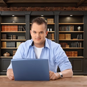 May include: A man in a light blue button-down shirt and a white undershirt sits at a wooden desk, holding a blue laptop. Behind him is a dark wooden bookcase filled with books, with a brick wall in the background.