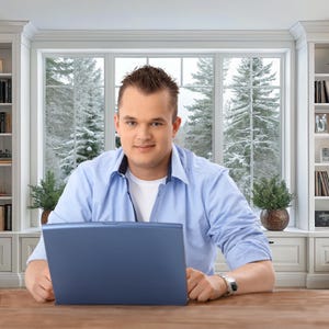 May include: A man wearing a blue button-down shirt and a white undershirt is seated at a wooden desk, holding a blue laptop. The background features a large window with a snowy outdoor scene and built-in white bookshelves.