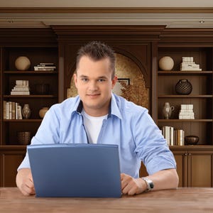 May include: A person wearing a blue button-down shirt and a white undershirt, sitting at a wooden desk. A blue laptop is open in front of them. The background features a dark wood bookcase filled with books and decorative items.