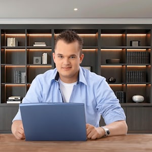 May include: A man in a light blue button-down shirt sits at a wooden desk, holding a blue laptop. A bookshelf with books and decorative items is in the background. The man is wearing a silver watch.