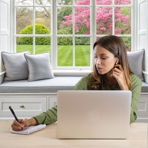 May include: A woman sits at a desk in front of a window with a view of a green lawn and a pink flowering tree. She is wearing a green shirt and is writing in a notebook with a black pen. A laptop computer is open in front of her.