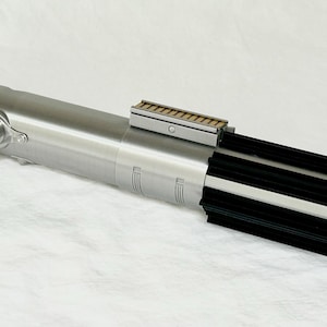 May include: A metallic silver lightsaber prop with black and silver details. The prop features a red button, a rectangular section with a series of small, parallel lines, and a black ribbed section at the end. The lightsaber is a replica of a popular science fiction weapon.