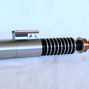 May include: A silver lightsaber with a black and silver handle, a copper coloured connector, and a black and silver emitter. The lightsaber has a small control panel with red, green, and white buttons.
