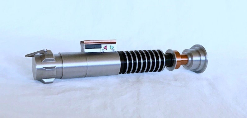 May include: A silver lightsaber with a black and silver handle, a copper coloured connector, and a black and silver emitter. The lightsaber has a small control panel with red, green, and white buttons.