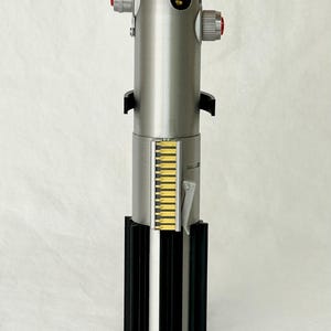May include: A metallic silver lightsaber prop with red and yellow accents. The base is black with a blue nameplate that reads "LUKE SKYWALKER". The lightsaber has a detailed design with various buttons and panels.