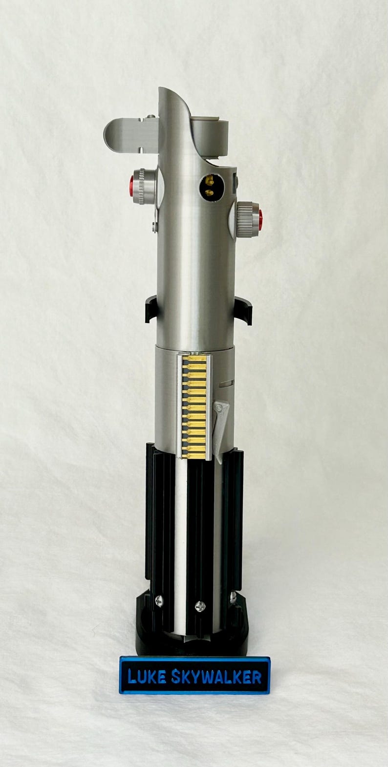 May include: A metallic silver lightsaber prop with red and yellow accents. The base is black with a blue nameplate that reads "LUKE SKYWALKER". The lightsaber has a detailed design with various buttons and panels.