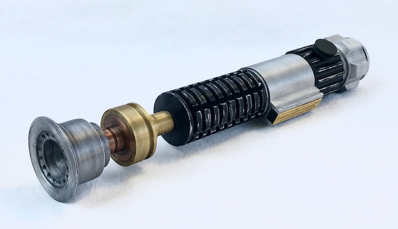 May include: A disassembled lightsaber with a black and silver handle, a gold and copper connector, and a silver emitter. The lightsaber is on a white surface.