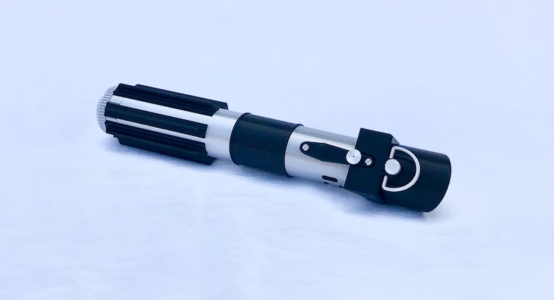 May include: A black and silver lightsaber prop with a black handle and a silver blade. The handle has a black grip and a silver metal band with a small black button.