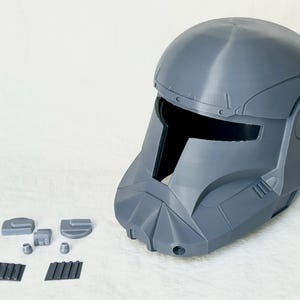 May include: A gray, 3D-printed helmet with a T-shaped visor and various detached components. The helmet has a rounded top and angular features, suggesting a futuristic or sci-fi theme. The detached pieces include small rectangular and square shapes.