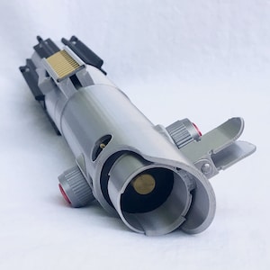 Puede incluir: Un accesorio de sable de luz plateado y gris con detalles en rojo y dorado. El accesorio presenta un dise&ntilde;o detallado con varios botones, interruptores y un emisor de hoja. El sable de luz es una r&eacute;plica de un arma de ciencia ficci&oacute;n popular.