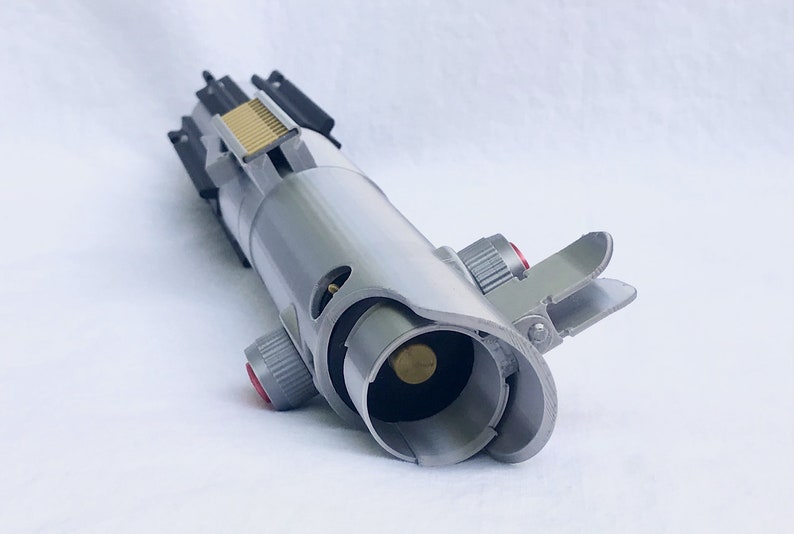 Puede incluir: Un accesorio de sable de luz plateado y gris con detalles en rojo y dorado. El accesorio presenta un dise&ntilde;o detallado con varios botones, interruptores y un emisor de hoja. El sable de luz es una r&eacute;plica de un arma de ciencia ficci&oacute;n popular.