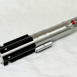 May include: A silver and black lightsaber prop with a red button and detailed metallic accents. The prop features a cylindrical body with black grips and a silver emitter, set against a white background.