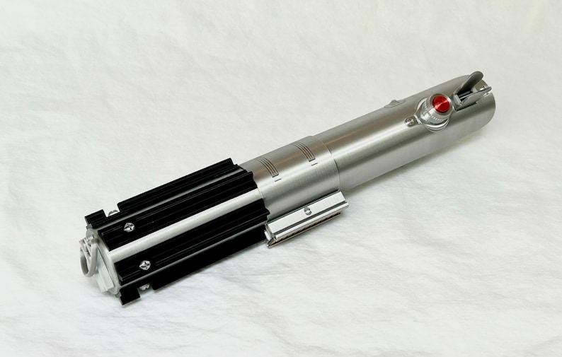 May include: A silver and black lightsaber prop with a red button and detailed metallic accents. The prop features a cylindrical body with black grips and a silver emitter, set against a white background.