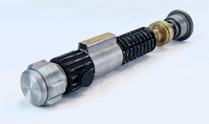 May include: A silver and black lightsaber prop with a brass accent. The lightsaber is made of metal and has a textured surface.