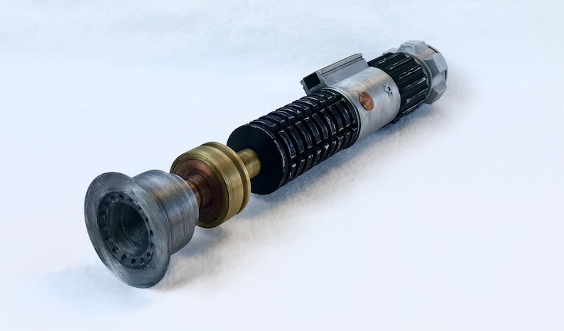 May include: A disassembled lightsaber with a black, silver, and gold hilt. The lightsaber is made of metal and has a detailed design. The lightsaber is on a white surface.