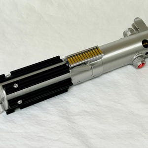 May include: A silver and black lightsaber prop with a detailed design. The hilt features a series of black fins, a silver handle, and a red button. The prop is a replica of a lightsaber from a popular science fiction franchise.