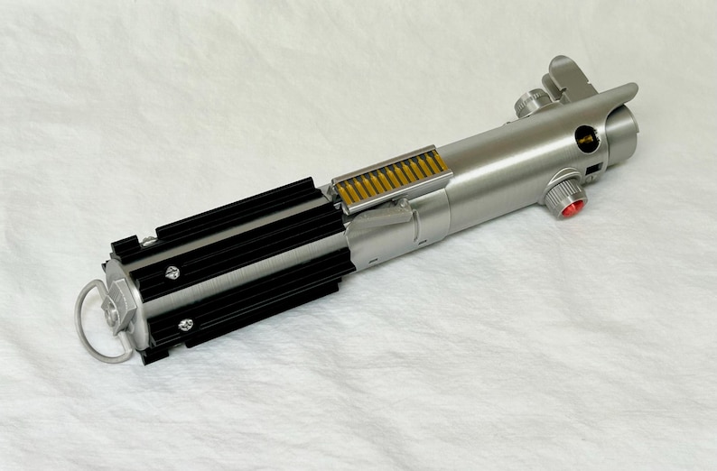 May include: A silver and black lightsaber prop with a detailed design. The hilt features a series of black fins, a silver handle, and a red button. The prop is a replica of a lightsaber from a popular science fiction franchise.