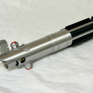 May include: A silver lightsaber prop with black and green details. The prop features a cylindrical body with a ribbed design, a black emitter, and red buttons. The lightsaber is a replica of a popular science fiction weapon.