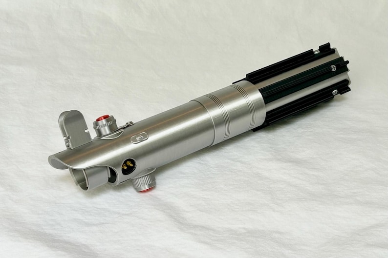 May include: A silver lightsaber prop with black and green details. The prop features a cylindrical body with a ribbed design, a black emitter, and red buttons. The lightsaber is a replica of a popular science fiction weapon.