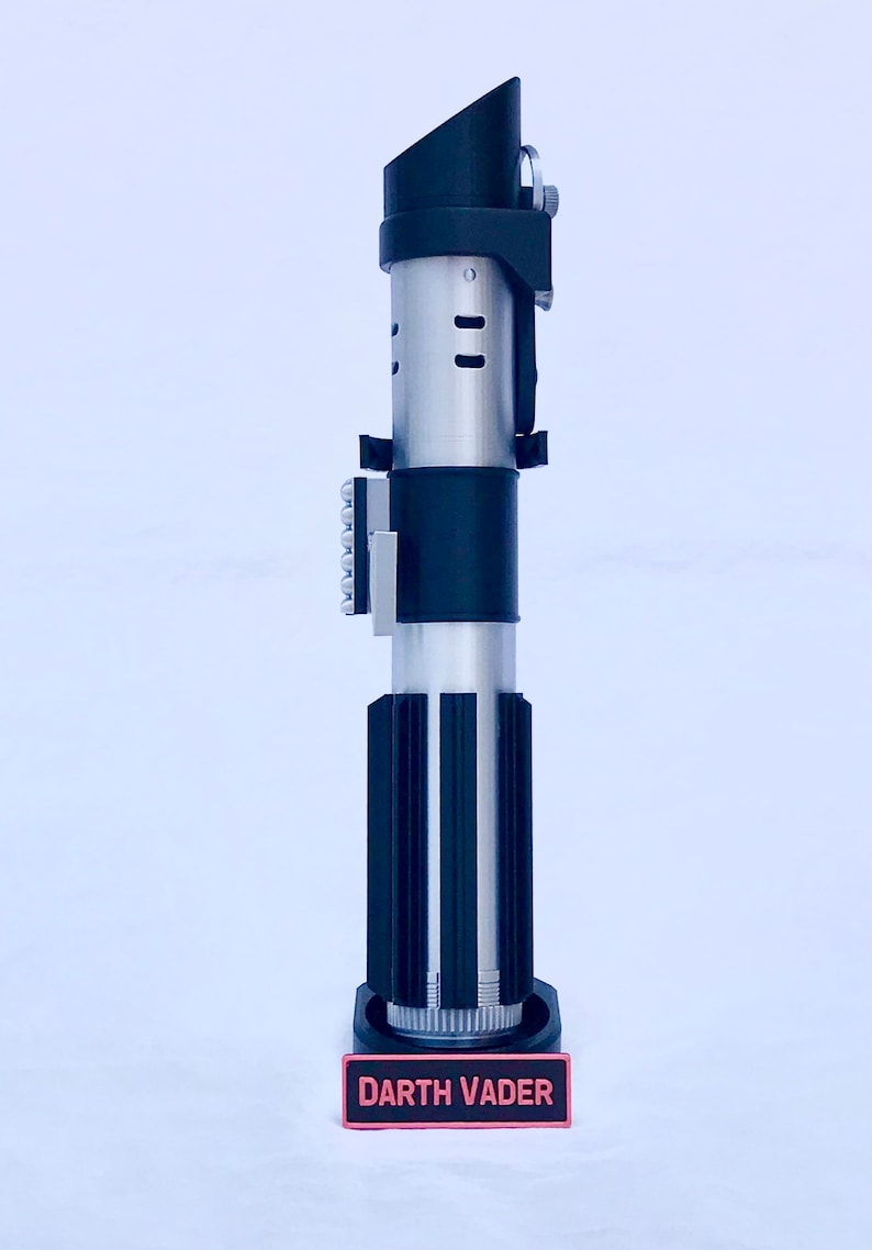May include: A black and silver lightsaber replica with a black stand. The stand has a red label that reads "DARTH VADER".