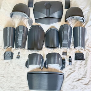 May include: A collection of gray 3D-printed armor pieces, including a chest plate, helmet sections, shoulder guards, and arm pieces. The components are arranged on a white surface, showcasing the various parts of the armor set.