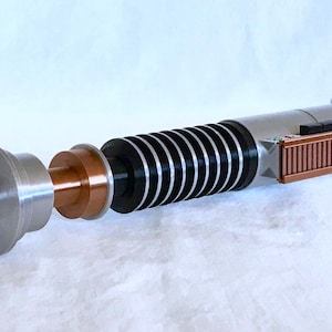 May include: A disassembled lightsaber with a silver, black, and copper coloured hilt. The lightsaber is made up of several pieces, including a cylindrical handle, a copper coloured ring, and a black and silver section with a series of black rings.