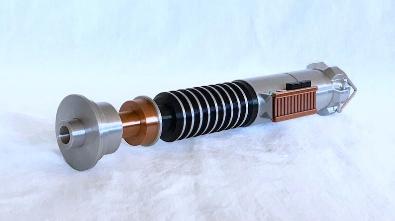 May include: A disassembled lightsaber with a silver, black, and copper coloured hilt. The lightsaber is made up of several pieces, including a cylindrical handle, a copper coloured ring, and a black and silver section with a series of black rings.