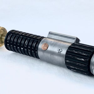 May include: A disassembled lightsaber with a silver and black body, a brass connector, and a silver cap. The lightsaber has a textured grip and a small copper coin on the side.