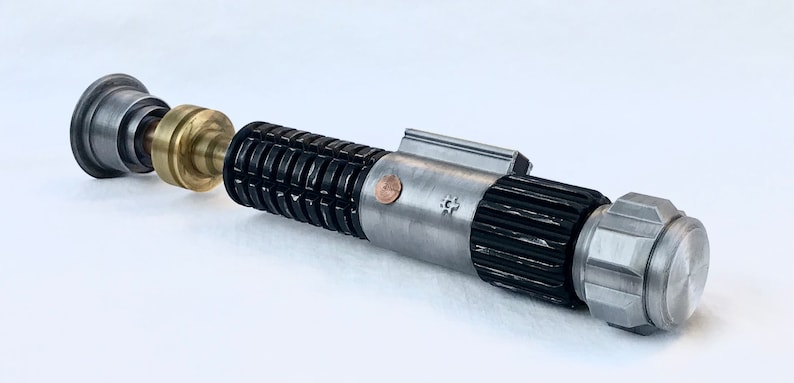 May include: A disassembled lightsaber with a silver and black body, a brass connector, and a silver cap. The lightsaber has a textured grip and a small copper coin on the side.