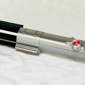 May include: A silver lightsaber prop with black and gold accents. The prop features a red button and a black switch. The lightsaber is lying on a white surface.