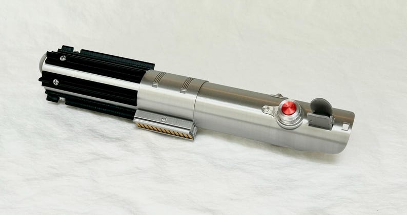 May include: A silver lightsaber prop with black and gold accents. The prop features a red button and a black switch. The lightsaber is lying on a white surface.