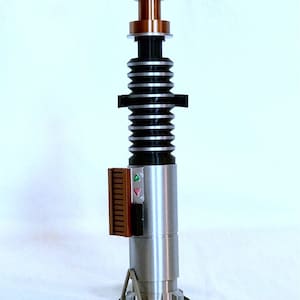 May include: A silver lightsaber with a black and silver handle, a brown and silver control panel, and a copper coloured emitter. The lightsaber is on a black stand with a green label that reads "LUKE SKYWALKER".