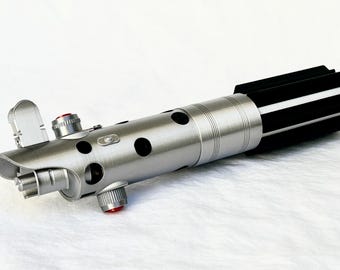 May include: A silver lightsaber hilt with a black and silver handle. The hilt has a red button and a silver lever.